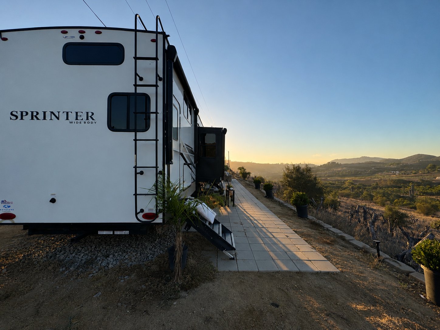 Exterior of The Golden Avocado fifth-wheel with paver walkway overlooking the hills at sunset